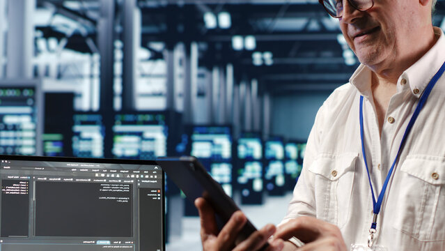 Senior technician in data center, solving tasks on tablet while running scripts on laptop terminal, with skilled technician amidst servers, accessing databases and manipulating lines of code.