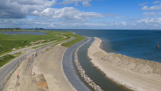 Aerial view of the dike reinforcement project along the coastline with a winding road, green embankments, and sandy construction works in Uitdam, North Holland, Netherlands.