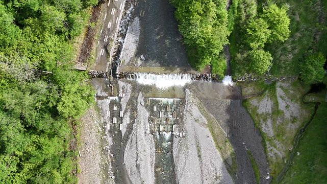 Aerial view of a river flowing through a forested valley. Water cascades over rocks, creating a small waterfall. Lush green trees surround the rocky streambed