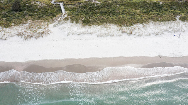 Aerial view of Omaha Beach with white sand, gentle waves, and a wooden walkway leading through coastal vegetation in Omaha, Auckland, New Zealand.