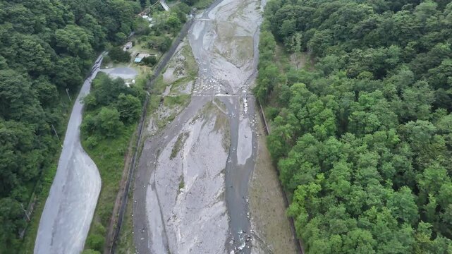 Aerial view of a dried riverbed. Trees flank the exposed riverbed. Grey sediment covers the dry channel. Road runs parallel to the riverbed. No people or animals visible