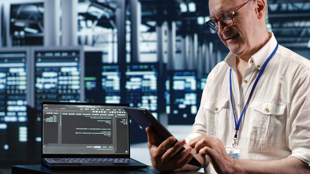 Elderly worker in data center, solving tasks on tablet while running scripts on laptop terminal, certified engineer amidst servers, accessing databases and manipulating lines of code.