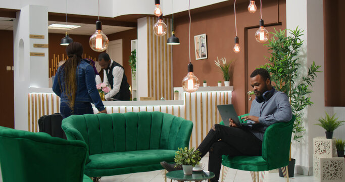 Traveller browsing social media at hotel, relaxing in lounge area with laptop before signing check in papers, African American person arriving at holiday resort, using computer on vacation.