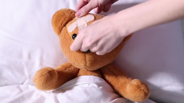Pediatrician concept. Doctor putting adhesive bandage onto toy bear in bed, closeup