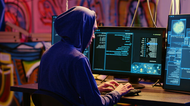 Hacker in graffiti painted hideaway base writing lines of code on computer, developing malware that get past security systems, rogue programmer uses PC in bunker, running hacking script.