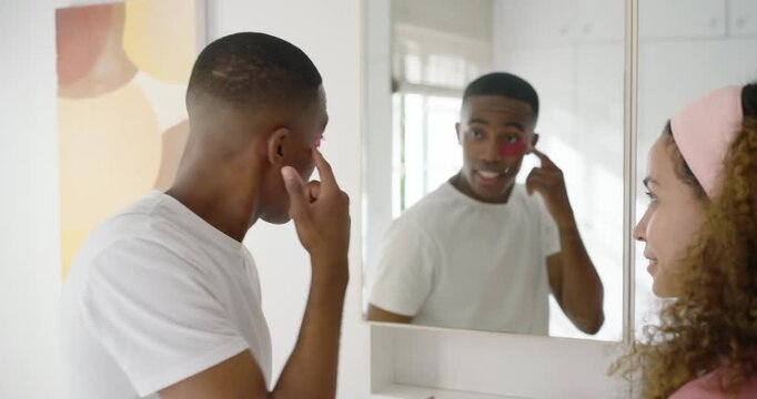 Couple at mirror, woman reaching, wearing headband applying pink eye patches for morning care