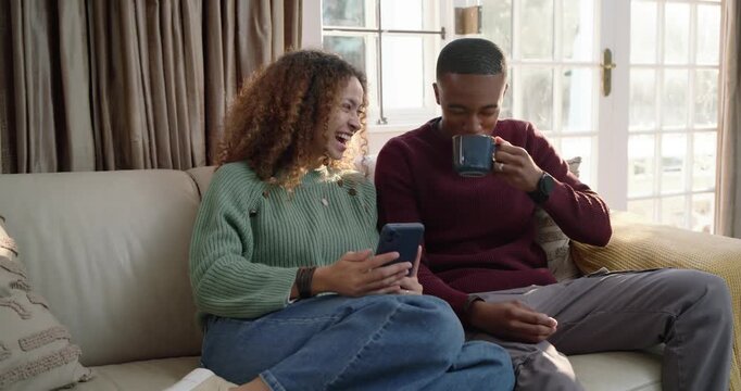 Diverse couple at home, woman showing phone sharing funny clip, man sipping blue mug laughing