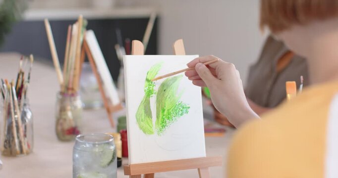 Female artists prompted by teacher, painting on easels applying green strokes while camera widening