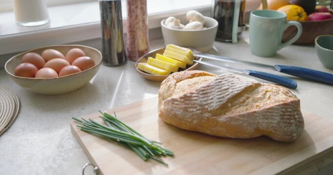 Camera is dollying by window, bringing crusty bread, butter and knives into focus for kitchen prep