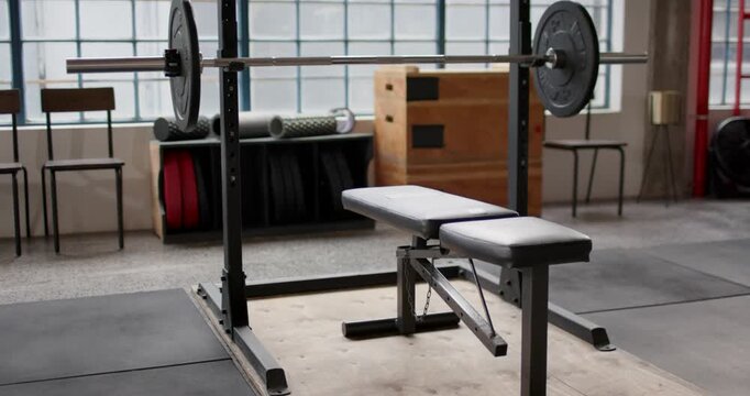 Cam is pulling back and panning upward revealing weight bench, loaded barbell, grid window, plates