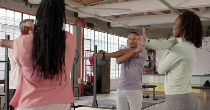Diverse group following right woman raising arm, warming up arms by power rack in warehouse gym