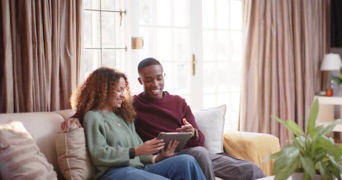 Couple sitting on living room sofa sharing tablet, woman tapping, man leaning and pointing, smiling