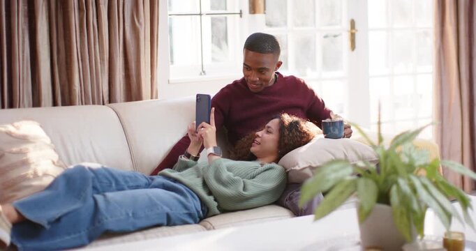 Diverse couple on sofa at home, woman scrolling phone, man leaning watching screen sipping blue mug