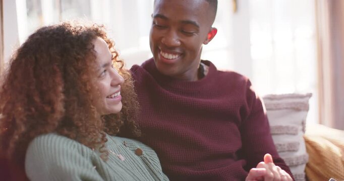 Diverse couple sitting on sofa, man in maroon knit holding hand, woman responding to touch, smiling