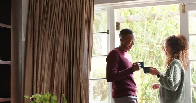 Diverse couple sharing brew by open casement-door with daylight streaming clinking mugs, copy space