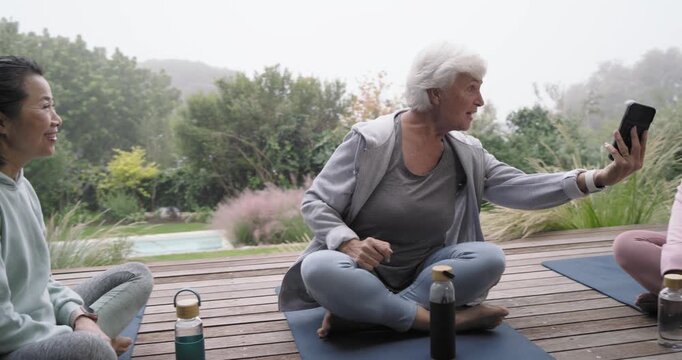 Diverse women in yoga attire on mats on deck, senior woman showing phone, sharing selfie, laughing
