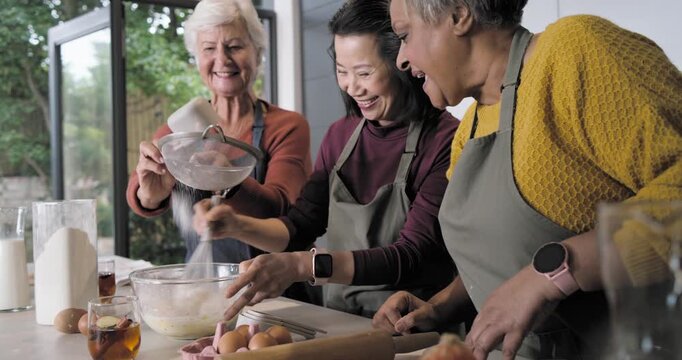 Diverse female friends in aprons sifting flour whisking batter pouring milk at counter for baking