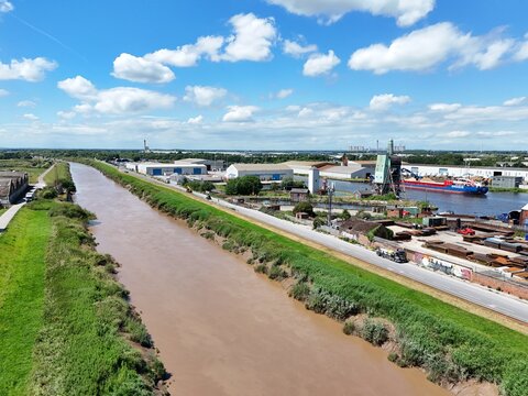 Aerial view of the canal in the industrial port area with cargo ships docked, warehouses, and green banks under a blue sky in Goole, England, United Kingdom.