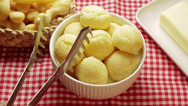 Authentic Brazilian cheese bread in a white ceramic bowl, with a plate of butter and a straw basket with cassava balls in the background
