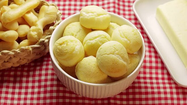 Authentic Brazilian cheese bread in a white ceramic bowl, with a plate of butter and a straw basket with cassava balls in the background. Brazilian breakfast.