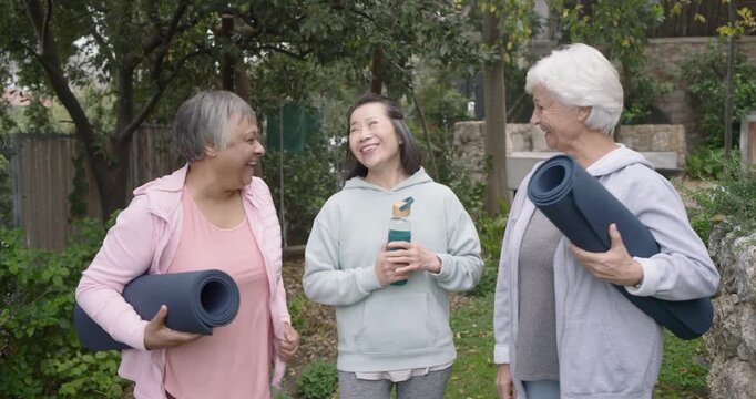 Diverse female friends arriving in garden, greeting, holding yoga mats and teal bottle for yoga