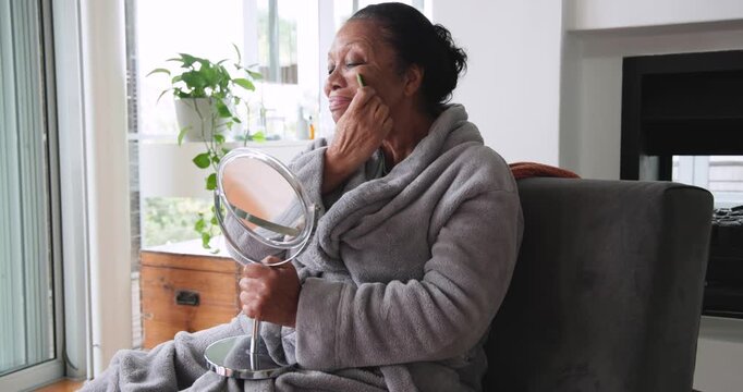 Senior African American woman sitting in robe at window holding mirror gliding stone for skincare