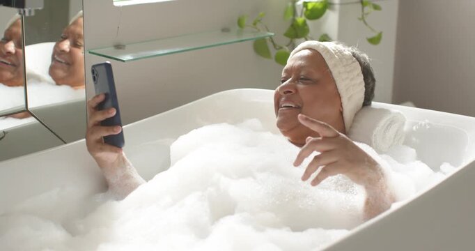African woman reclining in bubble bath, rolled towel, headband, holding phone, responding to phone