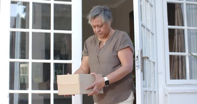 Senior woman wearing brown top receiving delivery holding cardboard box checking label for damage