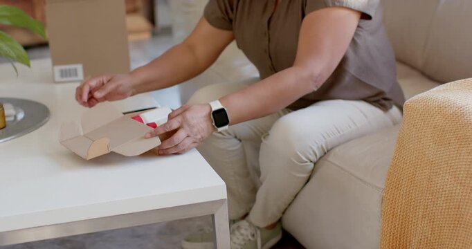 Senior African woman opening recent parcel on sofa, lifting bottle, checking phone, verifying label
