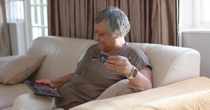 Senior woman sitting on sofa tapping tablet after payment prompt holding credit card for purchase