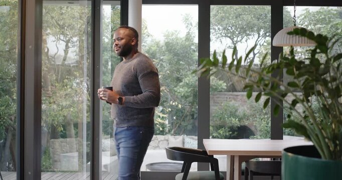 African American man entering with mug, moving to glass doors in dining area, gazing to reflect
