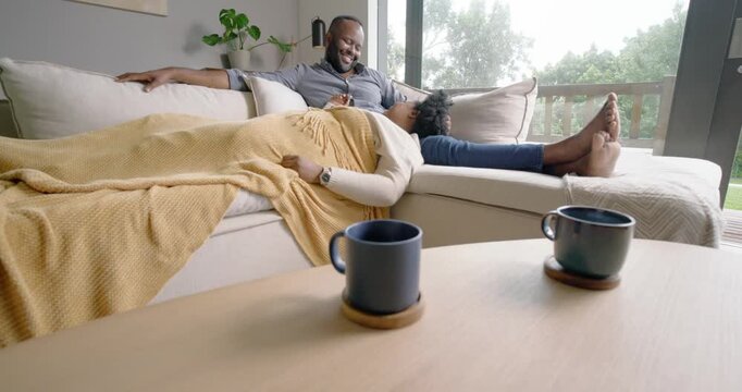 Couple relaxing on sofa by window with man smiling, looking down, using smartphone, patting blanket