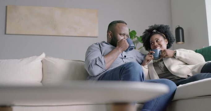 African American couple exchanging looks, sipping blue mugs, leaning on sectional sofa, connecting