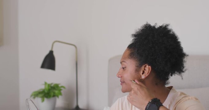 African American adult woman noticing blemish lifting hand mirror pressing cheek and checking skin