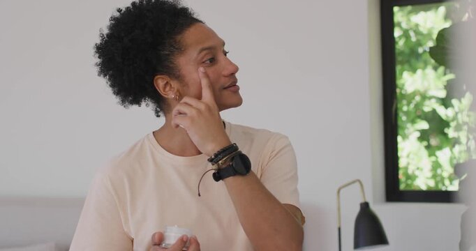 African American man taking cream from jar, applying to face, checking mirror, hydrating skin