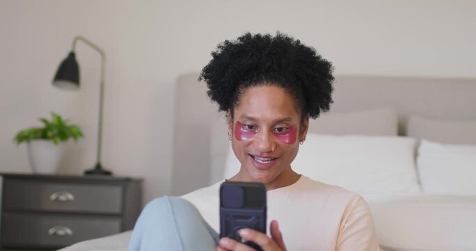 Adult African American woman smiling, waving at smartphone, tapping, checking pink eye patch on bed