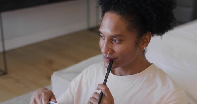 African American man studying, pausing pen at lips, then writing notes in spiral notebook on knee