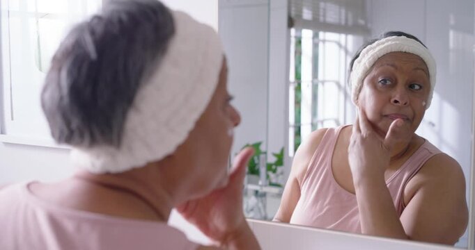 Senior African American woman with headband starting moisturizing at bathroom mirror, checking skin