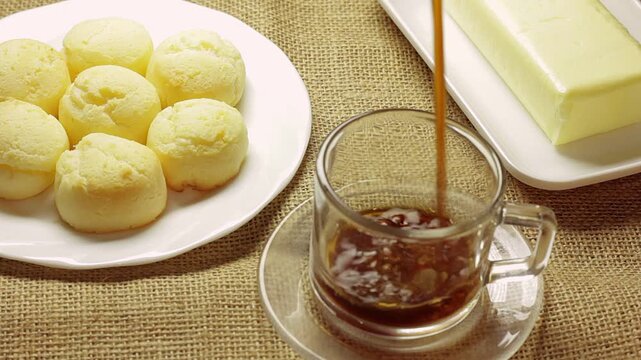 Authentic Brazilian cheese bread served with butter on a rustic burlap cloth, while fresh coffee is poured into a clear glass cup