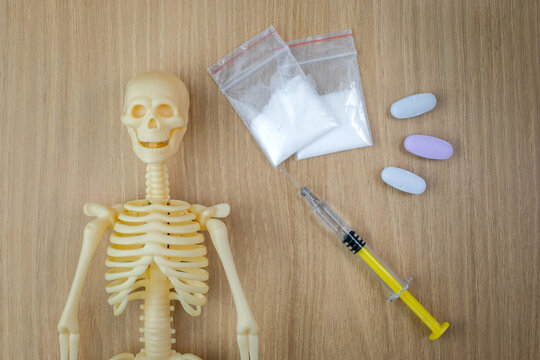 A skeleton anatomy model, syringe, tablets, and a small white powder bag are placed on a wooden table. Suitable for drug abuse prevention, overdose danger, addiction risk, or anti narcotics campaign