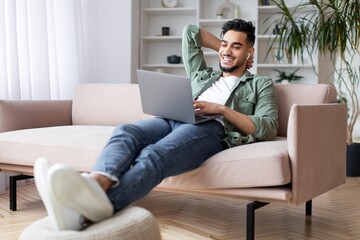 Obraz premium Smiling Young Man With Middle Eastern Descent Sitting Comfortably On Couch At Home, Using Laptop And Enjoying Music Through Earbuds, Creating A Relaxed Atmosphere In Living Room. Lifestyle Concept.