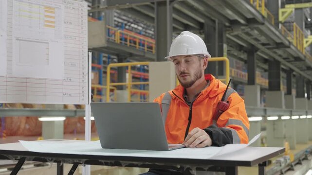Construction worker utilizes laptop to analyze blueprints at Train Depot Maintenance Depot, reviewing plans amidst industrial machinery and wearing white hard hat and orange safety vest.