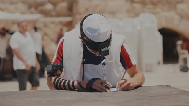 Jewish boy writing prayer at Western Wall Jerusalem wearing tefillin and tallit outdoors during daytime religious ceremony.