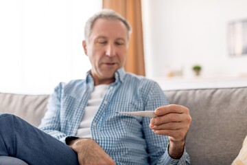 Fototapeta premium Worried Senior Caucasian Man Checking Body Temperature With Thermometer While Sitting On Couch At Home. Concerned About Health Condition. Healthcare Concept.