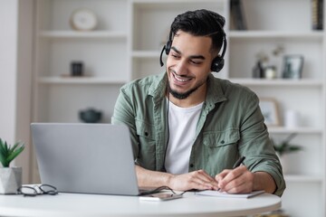 Obraz premium Smiling Young Hispanic Man Engaged In Remote Work With Laptop And Headphones In Cozy Home Office Setting. Focused On Tasks, Taking Notes, Representing Modern Business Concept.