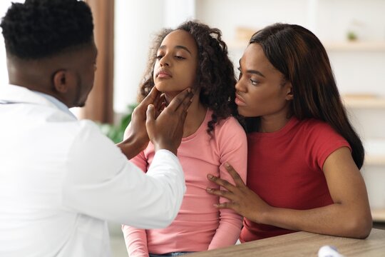 Worried Black Female Child About 8 Years Old Receiving Medical Examination From Doctor In Clinic While Her Mother Observes. Caring Interaction At Healthcare Office Setting.