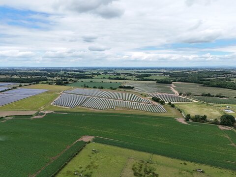 Aerial view of a large solar farm with rows of photovoltaic panels set in a green rural landscape under a cloudy sky in the United Kingdom.