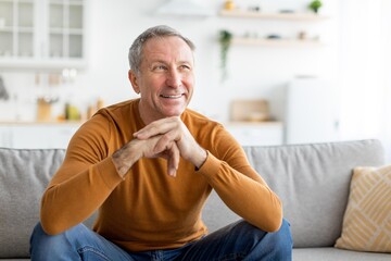 Fototapeta premium Smiling Elderly Caucasian Man Sitting Crosslegged On Couch With Relaxed Emotion At Home In Living Room, Enjoying Leisure Time. Concept Of Aging, Comfort, Relaxation And Lifestyle.
