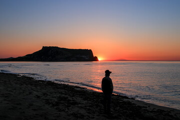 Fototapeta premium A beautiful sunrise over Seongsan Ilchulbong Peak in Jeju Island South Korea. A silhouette of a man stands on the quiet sandy beach looking at the golden sun rising next to the volcanic tuff cone.