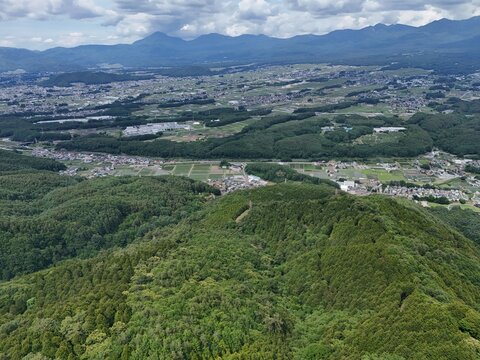 Green hills roll beneath a cloudy sky. Dense forests blanket the slopes below. Villages dot the valley, nestled among fields. Mountains loom far off, framing the horizon. Aerial view captures serene